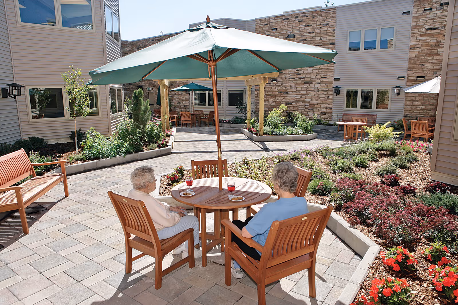 Two elderly women sitting at a wooden table with a green umbrella in a landscaped outdoor courtyard of a senior living facility. The courtyard features paved walkways, garden beds with flowers and shrubs, wooden benches, and the exterior walls of the building with windows.