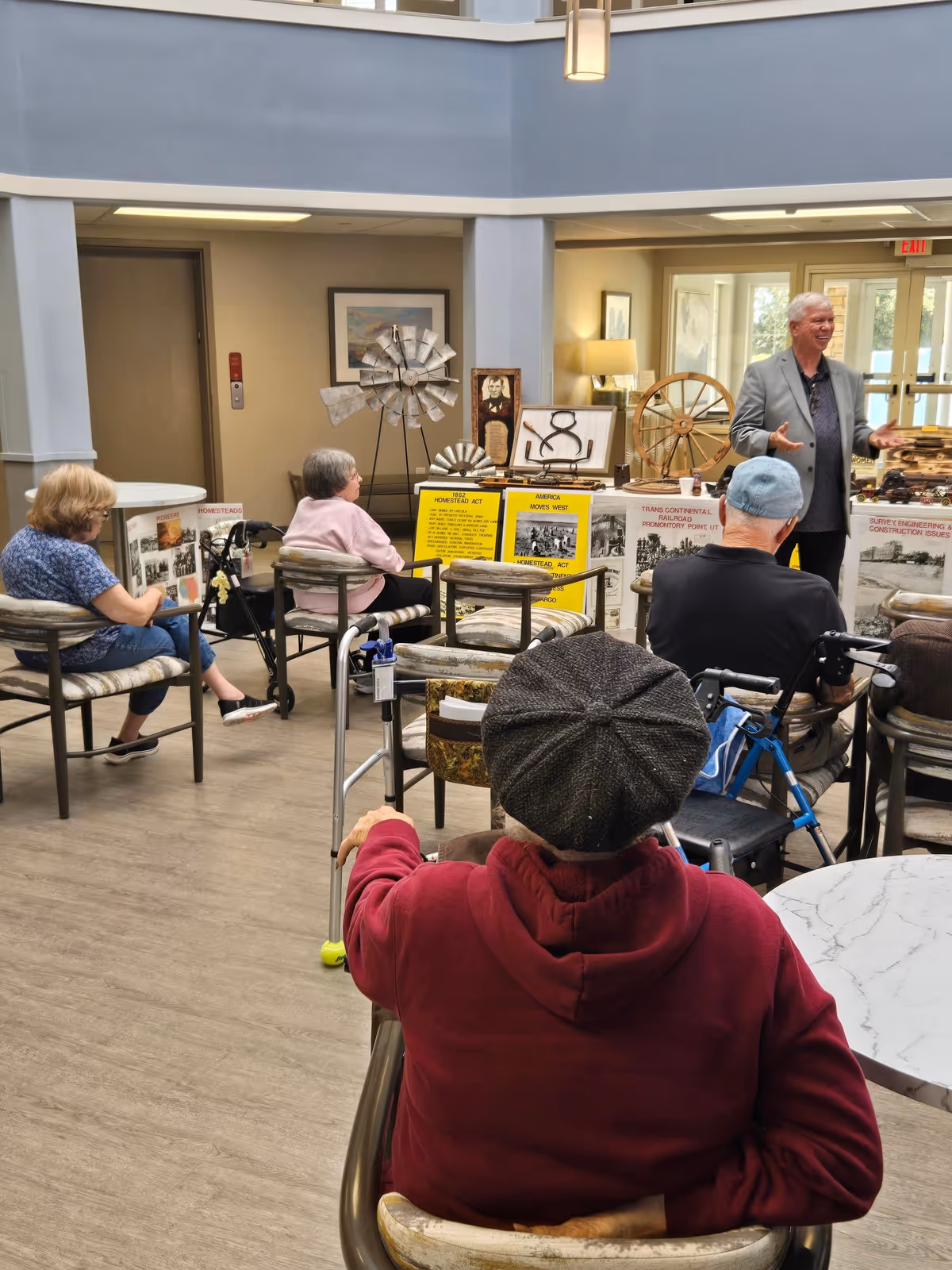 A group of elderly residents seated in a common room listening to a presenter standing before display boards and artifacts.