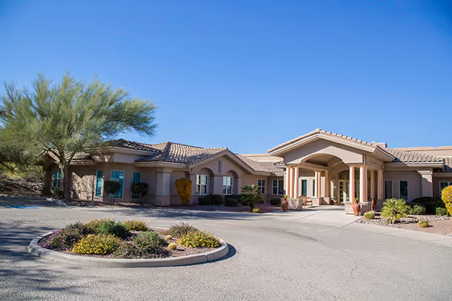 Exterior view of a single-story senior living facility building with a covered entrance, surrounded by desert landscaping including small bushes, trees, and rocks under a clear blue sky.