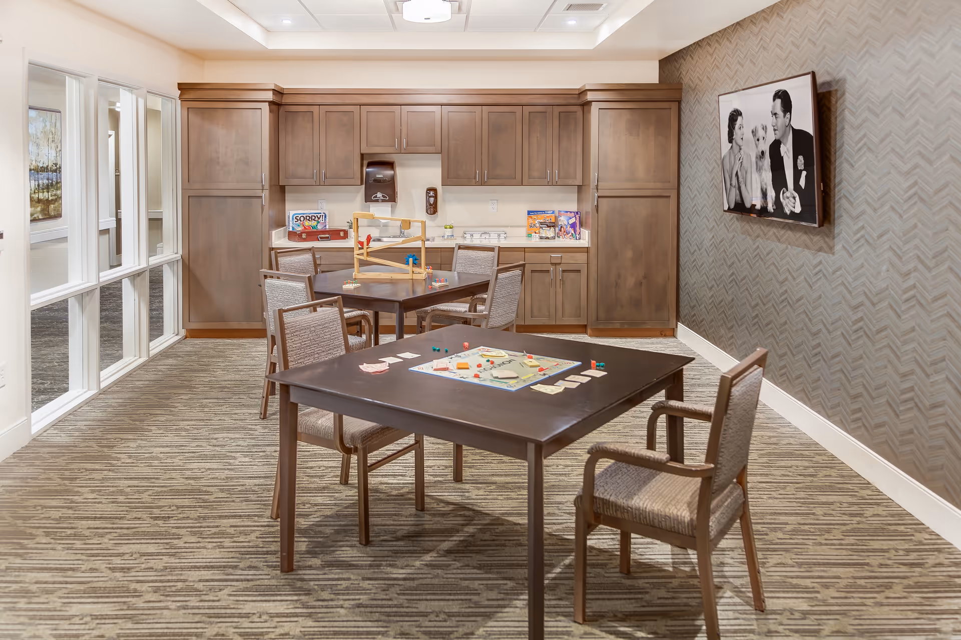 An interior activity room with tables and chairs, board games on the tables, and wooden cabinets along the back wall.
