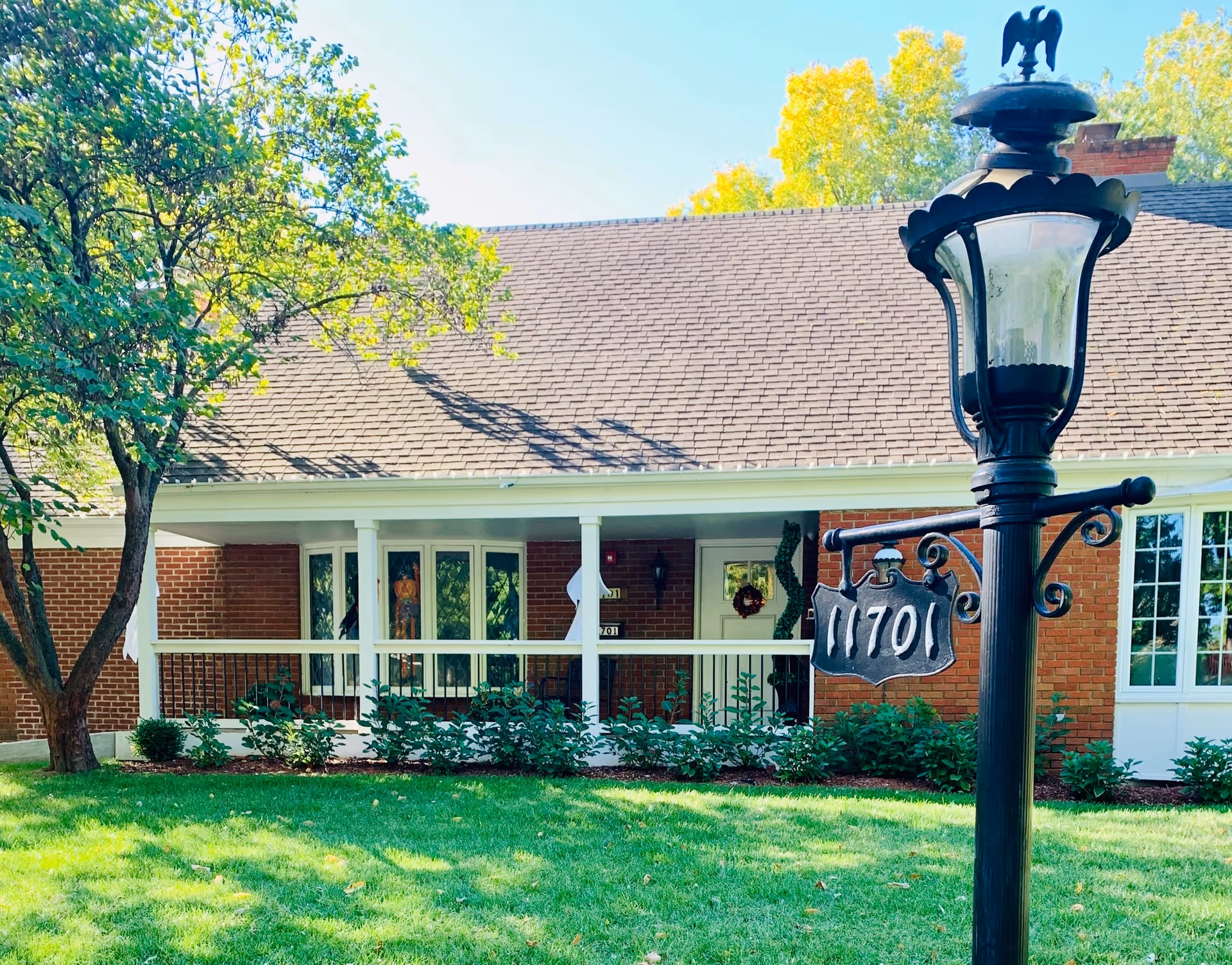 Front exterior view of a single-story brick building with a porch, white railings, and a green lawn. A black lamp post with the number 11701 is prominently visible in the foreground. Trees with green and yellow leaves surround the building.