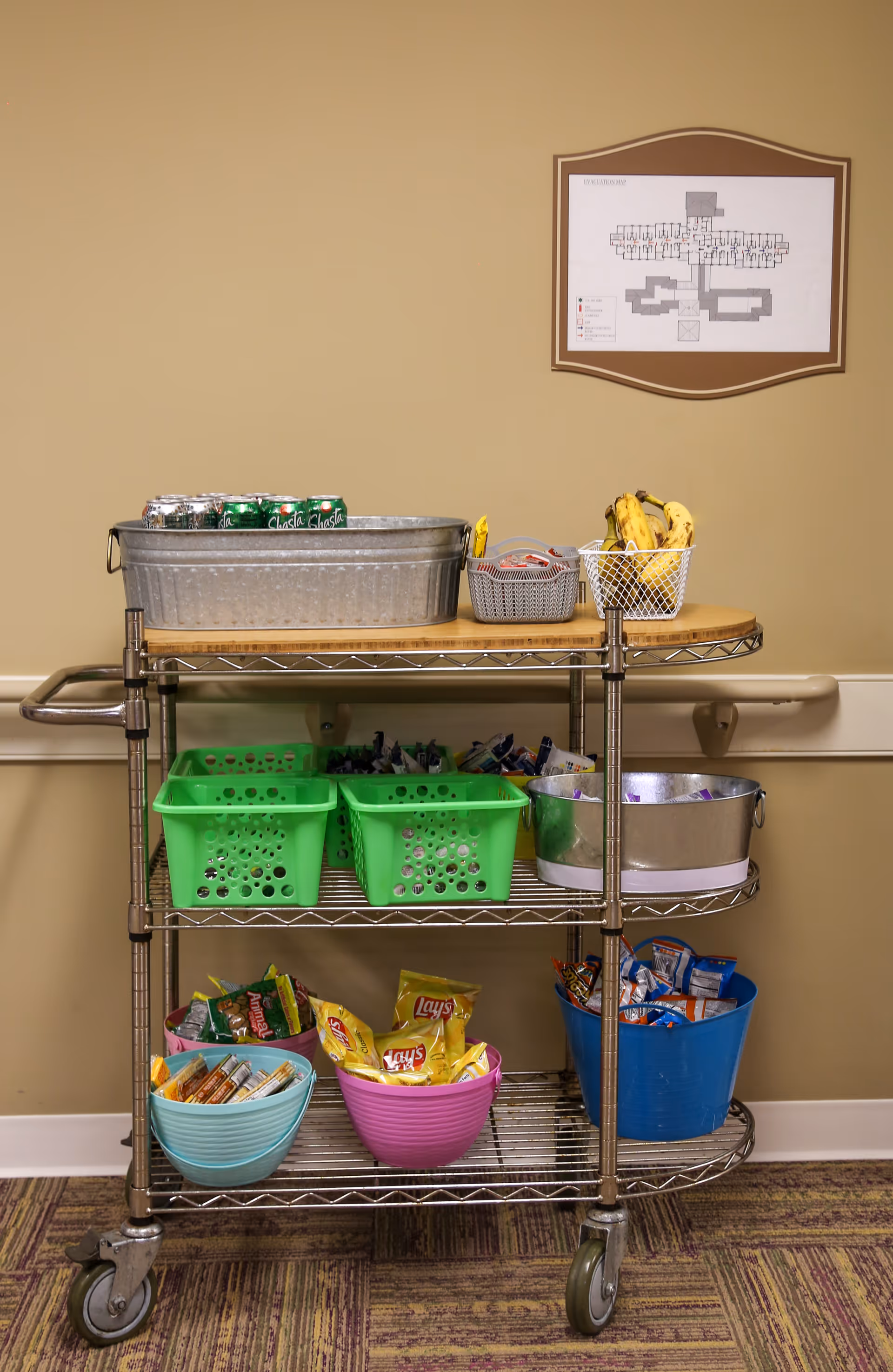 A metal rolling cart with three shelves holding various snacks and drinks. The top shelf has a metal tub with canned sodas, a small basket with packets, and a basket of bananas. The middle shelf has green plastic baskets and a metal tub with more snacks. The bottom shelf has colorful bowls filled with snack bags like Lay's chips and other assorted items. A floor plan is mounted on the beige wall behind the cart.