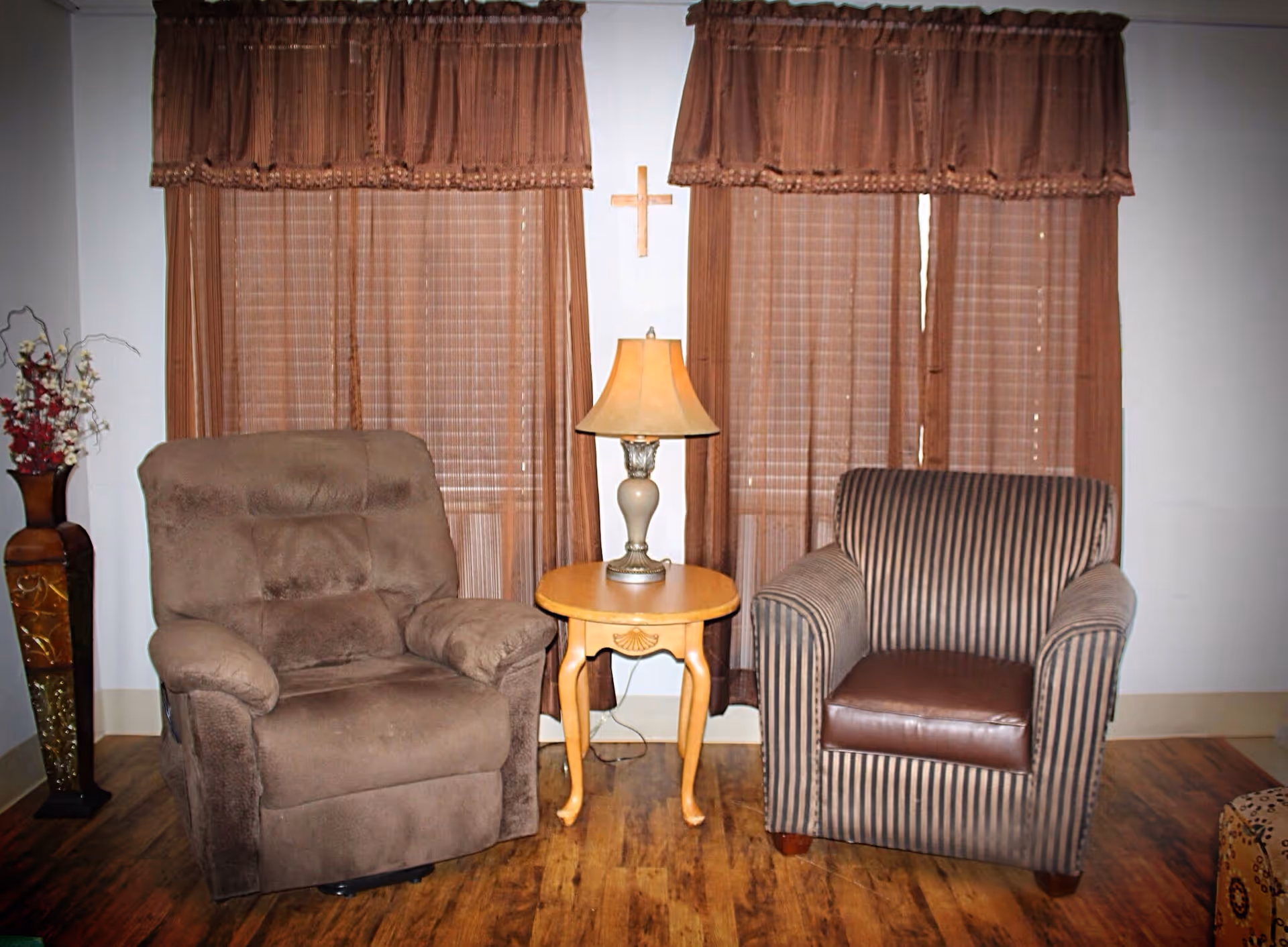Two armchairs flanking a small round table with a lamp in front of closed brown curtains and a wall cross.
