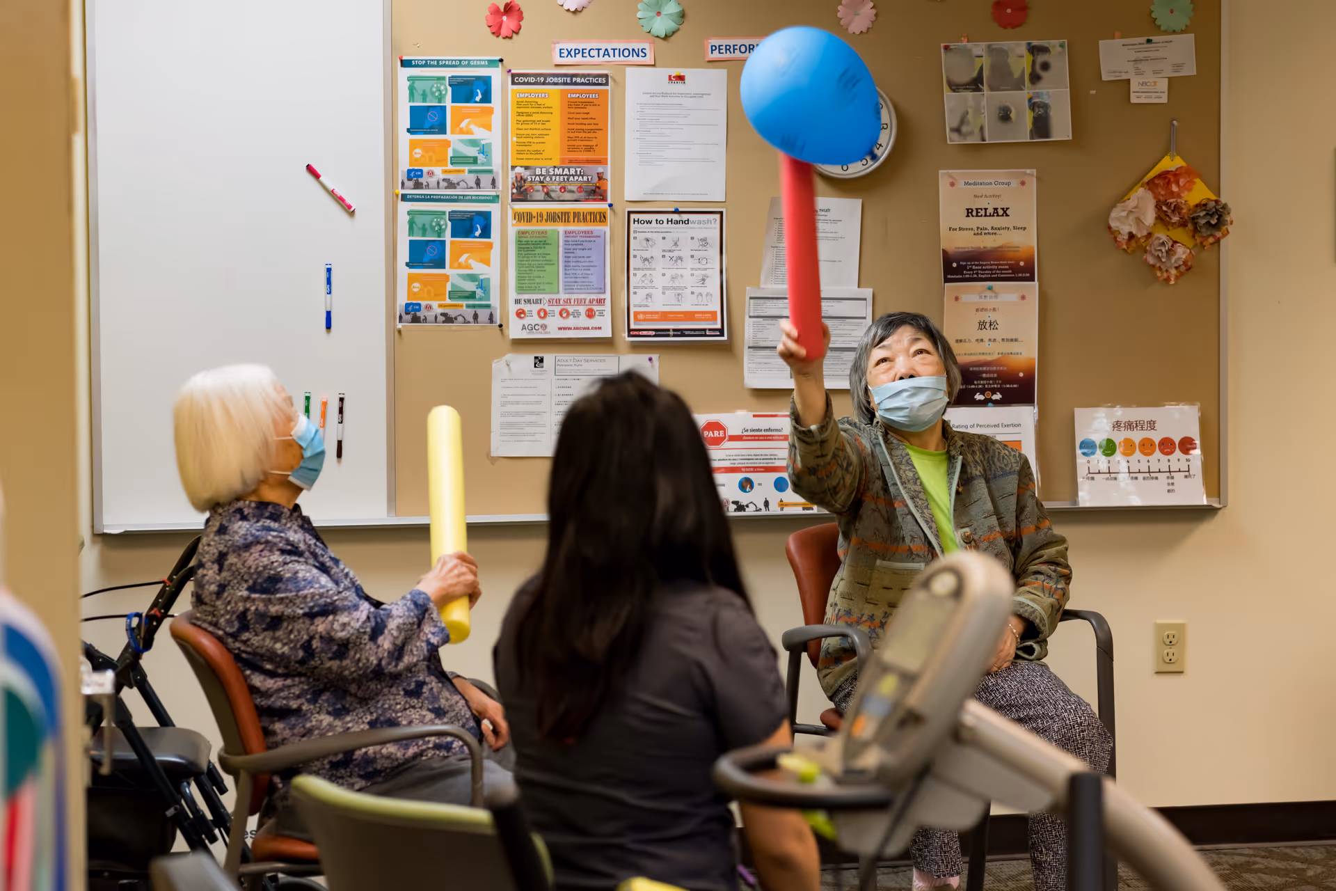 Three elderly women sitting in chairs in a room with a bulletin board covered in notices and posters. Two women are holding foam sticks, one yellow and one red with a blue balloon attached, and appear to be engaging in a light exercise or activity. All three women are wearing face masks.