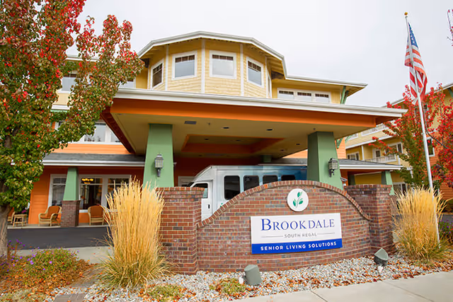 Exterior view of a senior living facility with a covered entrance supported by green pillars. There is a brick sign in front that reads 'Brookdale South Regal Senior Living Solutions'. The building has yellow and orange siding, multiple windows, and an American flag on a pole to the right. Trees with red autumn leaves and ornamental grasses surround the entrance area.