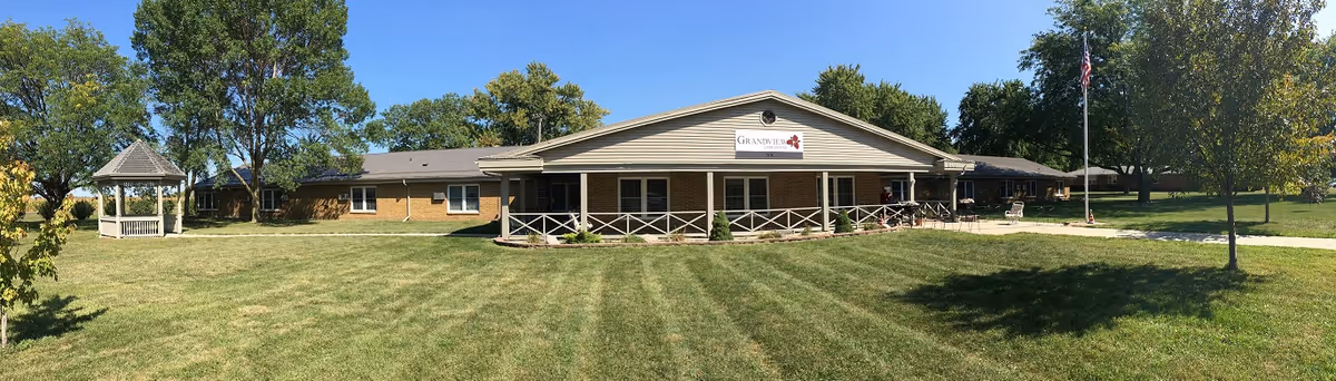Wide exterior view of Grandview Health Care Center, a single-story brick building with a covered porch and a sign displaying the facility's name. The building is surrounded by a well-maintained lawn with trees and a white gazebo on the left side. The sky is clear and blue.