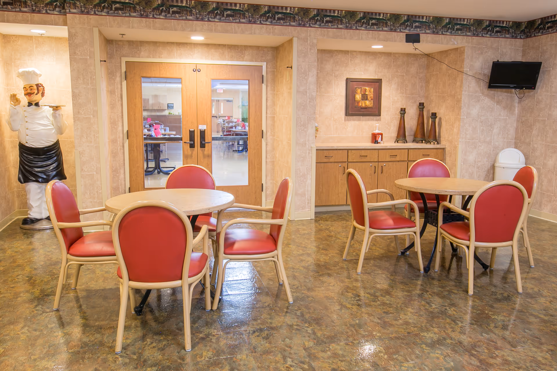 Interior room with two round tables each surrounded by four red cushioned chairs. A large chef statue holding a tray stands in the corner near double wooden doors with glass panels. A cabinet with decorative items and a small wall-mounted TV are visible on the right wall.