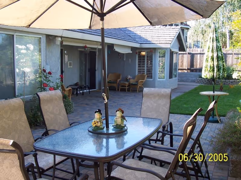Outdoor patio with a glass-top table and umbrella surrounded by chairs on a paved backyard in front of a single-story house.