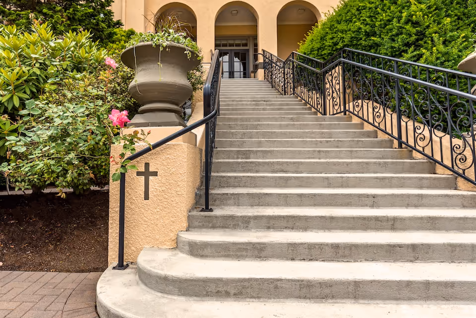 Wide concrete staircase with black wrought iron railings on both sides leading up to an entrance with three arches. There is a large planter with greenery on the left side and bushes on both sides of the stairs. A cross symbol is visible on the left wall near the base of the stairs.