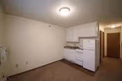Small kitchenette area with white cabinets, a white refrigerator, a white stove, and a sink. The room has beige walls and carpeted flooring. There is a hallway with a closed wooden door at the end.
