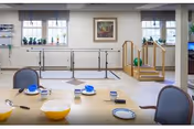 Bright multipurpose therapy room with parallel walking bars and exercise equipment, and a table set with bowls and cups in the foreground.
