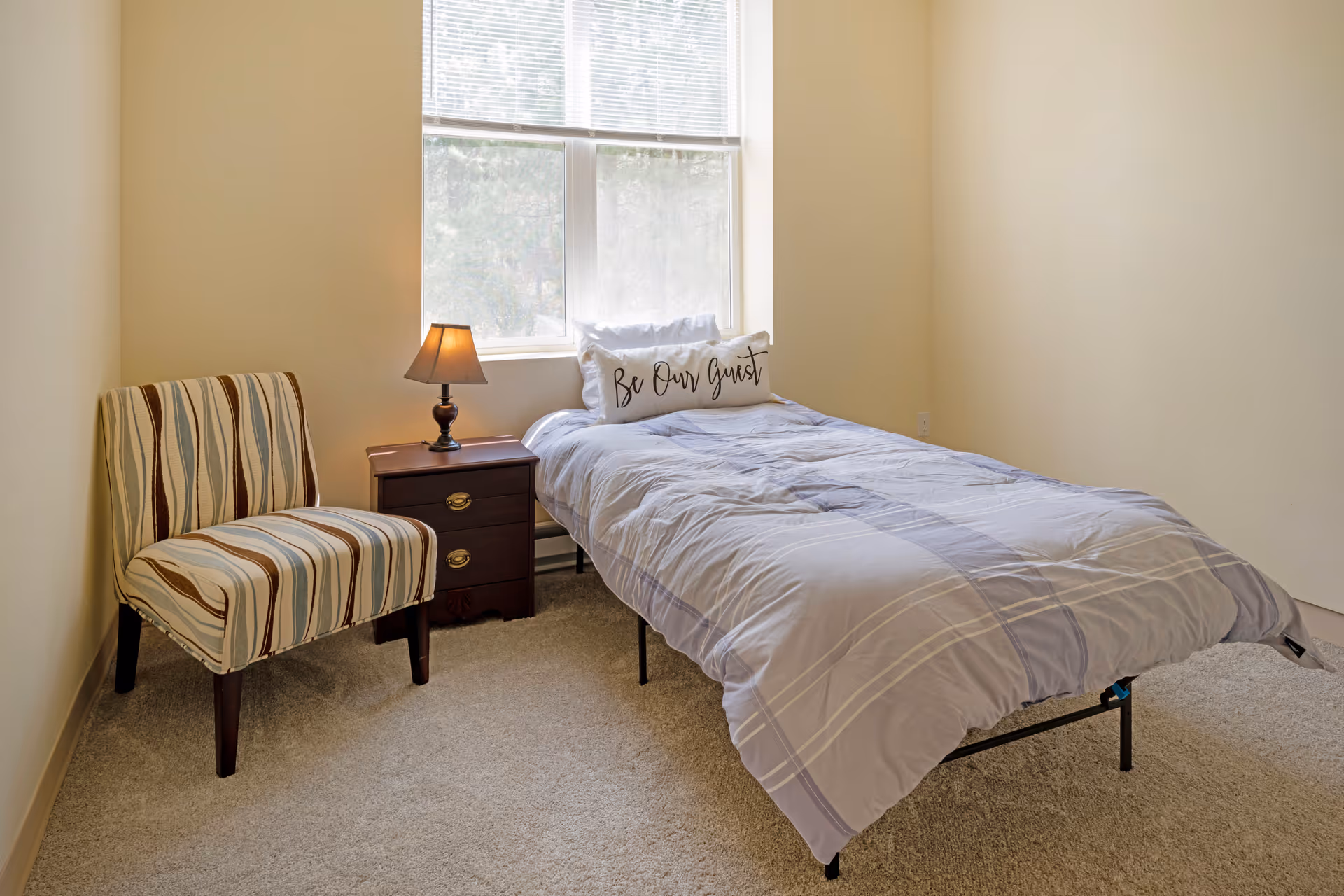 A small bedroom with a single bed covered in a light gray and white striped comforter, a pillow with the words 'Be Our Guest', a wooden nightstand with a lamp, and a striped upholstered chair. A large window with blinds lets in natural light.