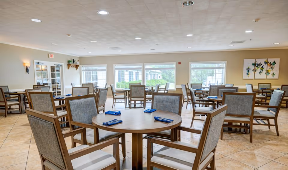 Spacious dining room with round wooden tables and upholstered chairs set with blue napkins and large windows.