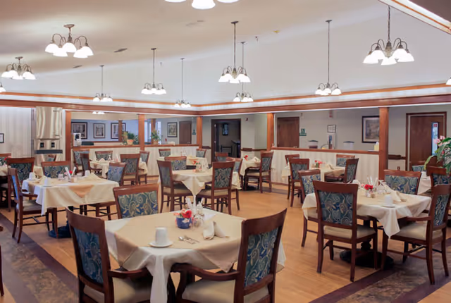 A bright, spacious dining room with multiple tables set with tablecloths and chairs under hanging light fixtures.