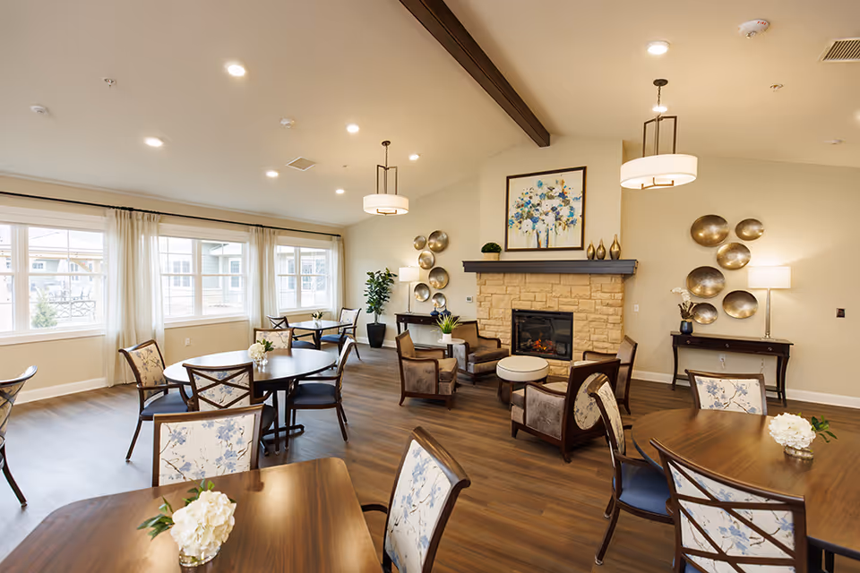 Well-lit communal dining and lounge area with round tables, upholstered chairs, and a stone fireplace.