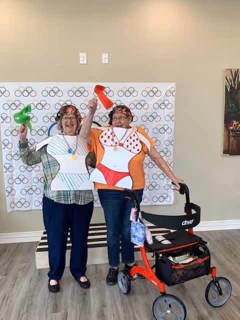 Two elderly women standing indoors in front of a backdrop with Olympic rings. They are wearing humorous paper cutouts of swimsuits over their clothes and holding colorful plastic megaphones. One woman is using a red walker. Both are smiling and appear to be enjoying themselves.