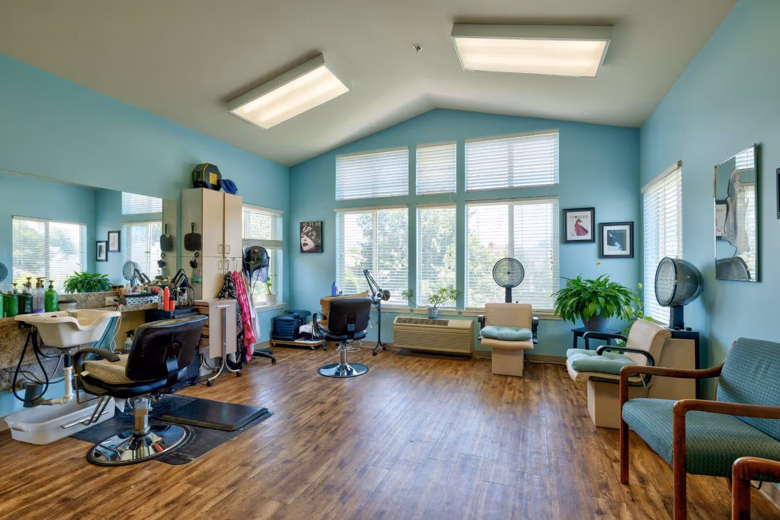 Bright salon-style room with hairdressing chairs, sinks, hood dryers and seating beneath large windows.