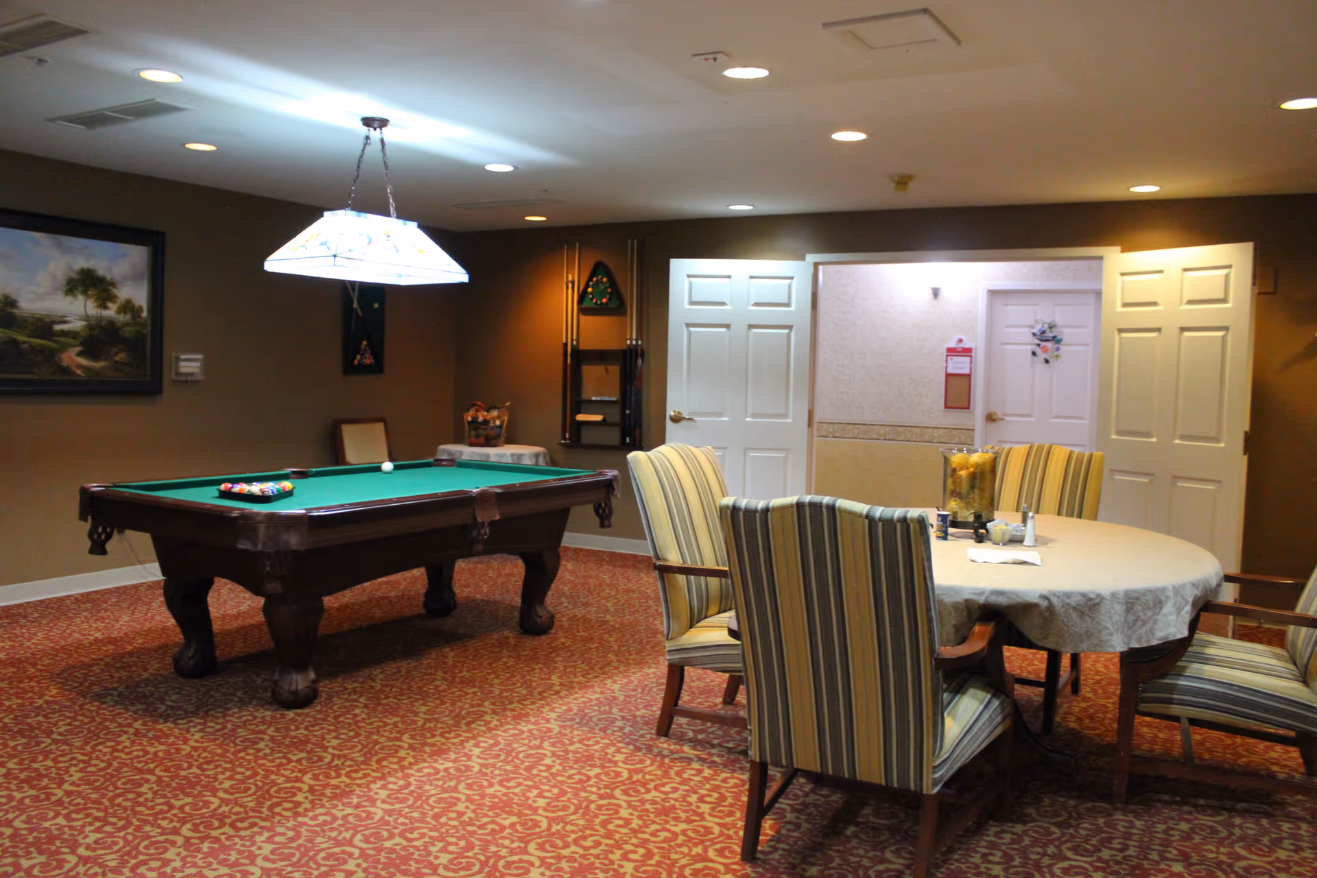 A senior living common room with a pool table, a round table surrounded by striped chairs, and doors in the background.