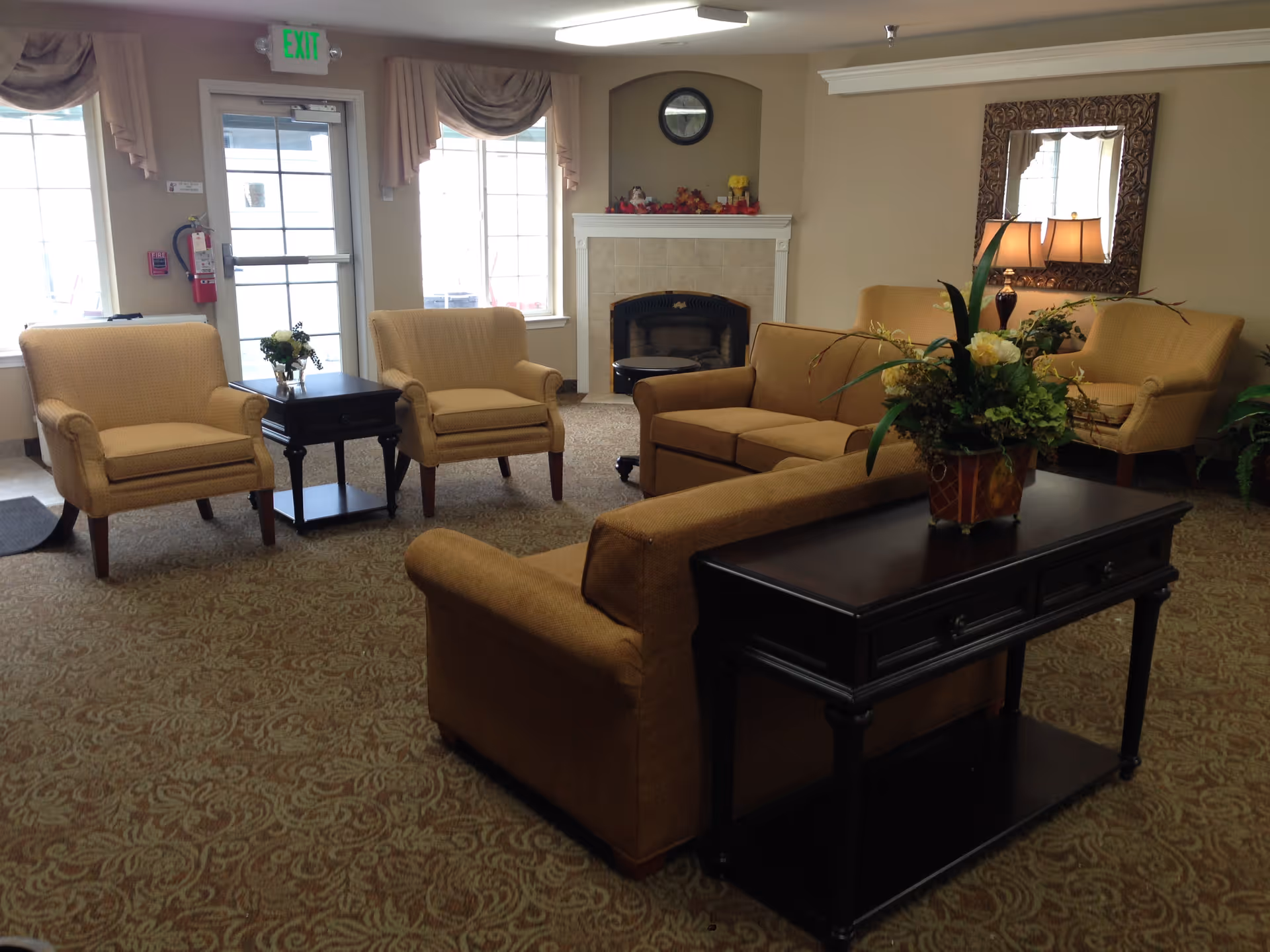 A cozy senior living facility common area with beige armchairs and sofas arranged around a dark wooden coffee table and side tables. There is a fireplace with a clock above it and decorative items on the mantel. A large mirror and a lamp are on the wall, and a floral arrangement is on the table behind the sofa. The room has carpeted flooring and windows letting in natural light.