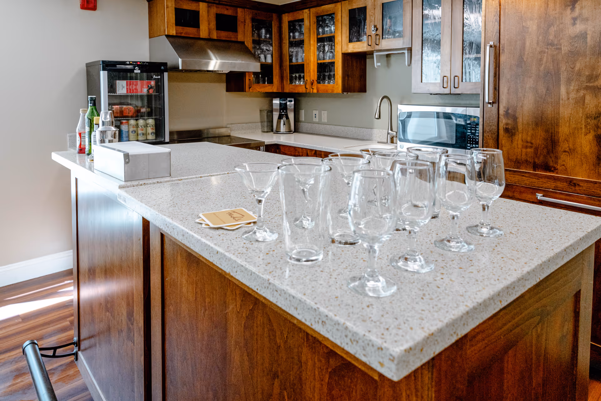 A kitchen area with a large island countertop featuring multiple empty glassware including wine glasses and a martini glass. The kitchen has wooden cabinets, a microwave, a coffee maker, a small refrigerator stocked with beverages, and a stainless steel range hood.