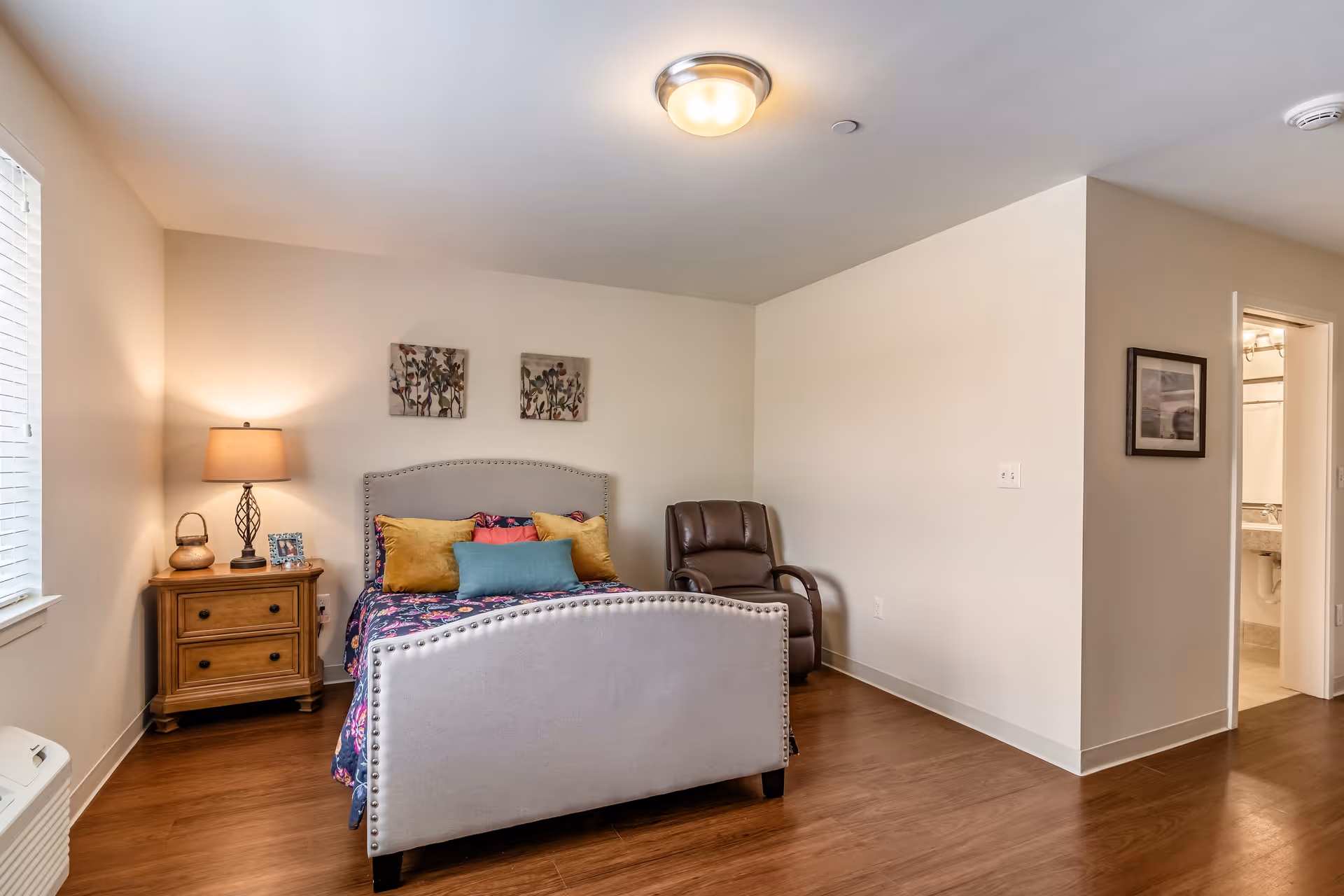 A cozy bedroom with a bed featuring a light gray upholstered headboard and footboard, decorated with colorful pillows and a floral bedspread. To the left of the bed is a wooden nightstand with a lamp, a small basket, and a framed photo. A brown leather recliner chair is placed in the corner to the right of the bed. The room has wooden flooring, beige walls, and a ceiling light fixture. A doorway on the right side leads to a bathroom with a sink and mirror visible.