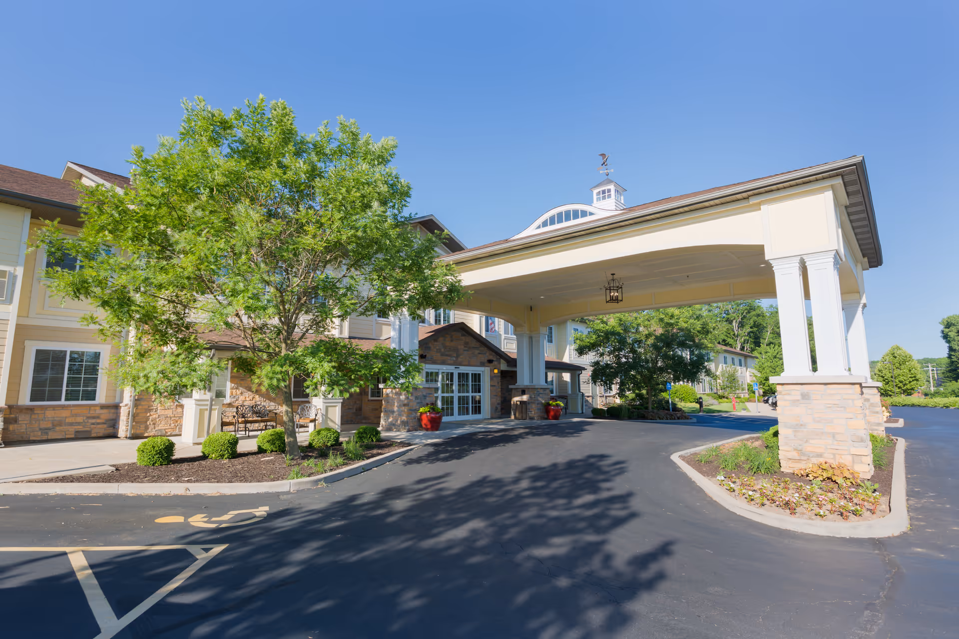 Entrance of Westview at Ellisville Assisted Living facility showing a covered drop-off area with columns, a tree, landscaped bushes, and a clear blue sky.