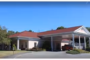 Exterior view of a single-story healthcare facility building with a covered entrance and a red roof under a clear blue sky. There is a driveway leading to the entrance and some greenery around the building.