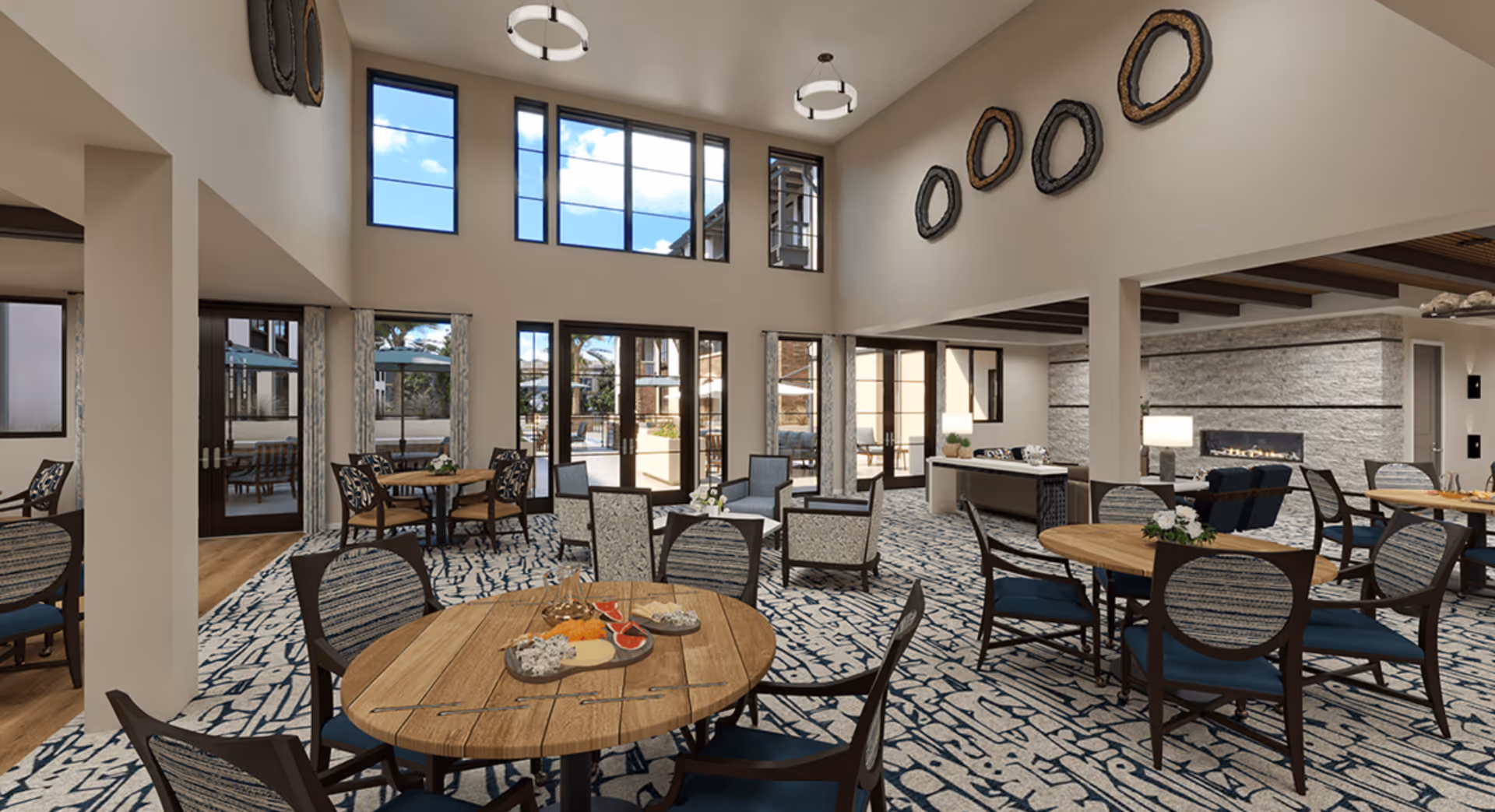 Spacious senior living common area with multiple round wooden tables and cushioned chairs arranged on a patterned carpet. Large windows and glass doors allow natural light to fill the room, with views of an outdoor patio. The room features high ceilings, modern light fixtures, decorative wall art, and a stone fireplace in the background.