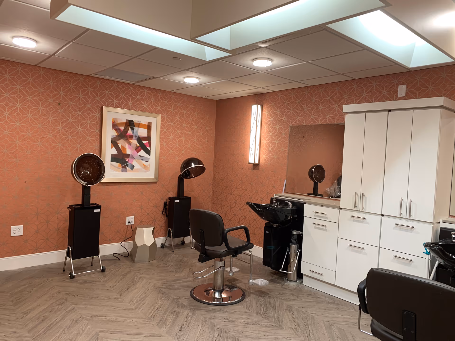 Interior of a hair salon area in a senior living facility with two black salon chairs, two hair drying stations, a white cabinet with drawers, a large mirror, and a framed abstract artwork on a patterned orange wall.