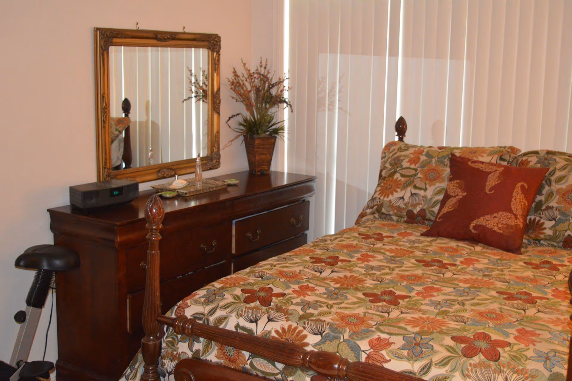 A bedroom with a wooden bed frame and floral patterned bedding in shades of orange, green, and beige. There is a matching pillow with a paisley design. Next to the bed is a dark wooden dresser with a decorative mirror above it. On the dresser are a small clock radio, a tray with a bottle and other small items, and a vase with artificial flowers. Vertical blinds cover the window behind the bed.