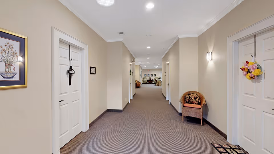 A well-lit hallway in an assisted living facility with beige walls and carpeted floor. Several white doors line both sides of the hallway, some decorated with hanging ornaments and wreaths. A wicker chair with a cushion is placed against the right wall. At the end of the hallway, a seating area with chairs and framed pictures on the wall is visible.