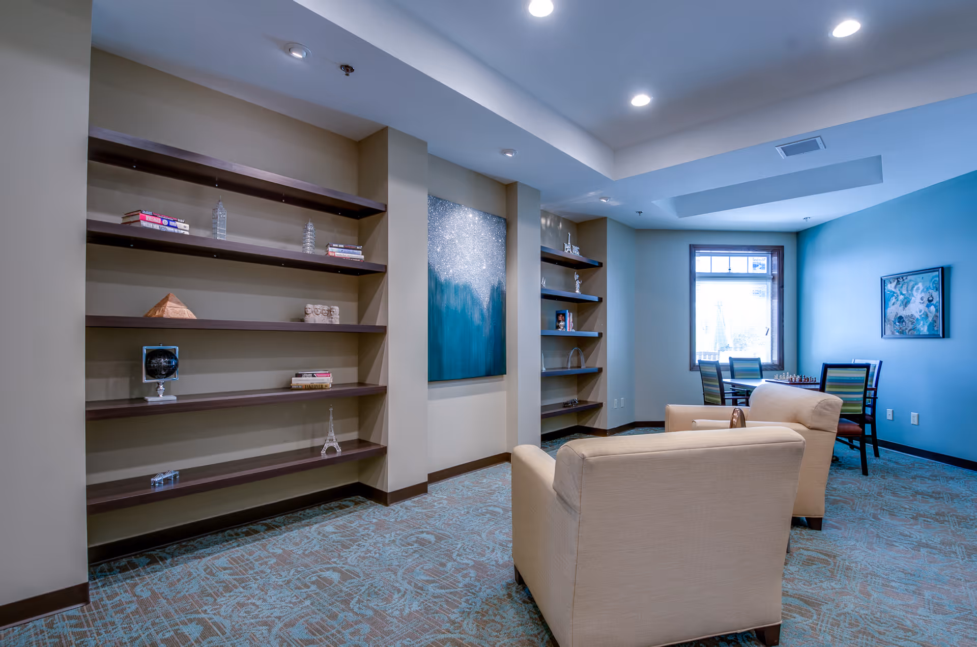 A cozy lounge area with two beige armchairs facing a small table. The room features built-in dark wooden shelves with decorative items and books, a large abstract painting on the wall, a window letting in natural light, and a small table with four chairs in the corner. The walls are painted in neutral tones with one accent wall in blue, and the floor is carpeted with a patterned design.