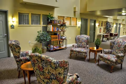 A cozy indoor seating area in an assisted living facility featuring four floral upholstered armchairs arranged around two small wooden tables. The space has beige walls with several doors and windows, a carpeted floor, and a metal bookshelf with books and decorative items. Potted plants add greenery to the area.