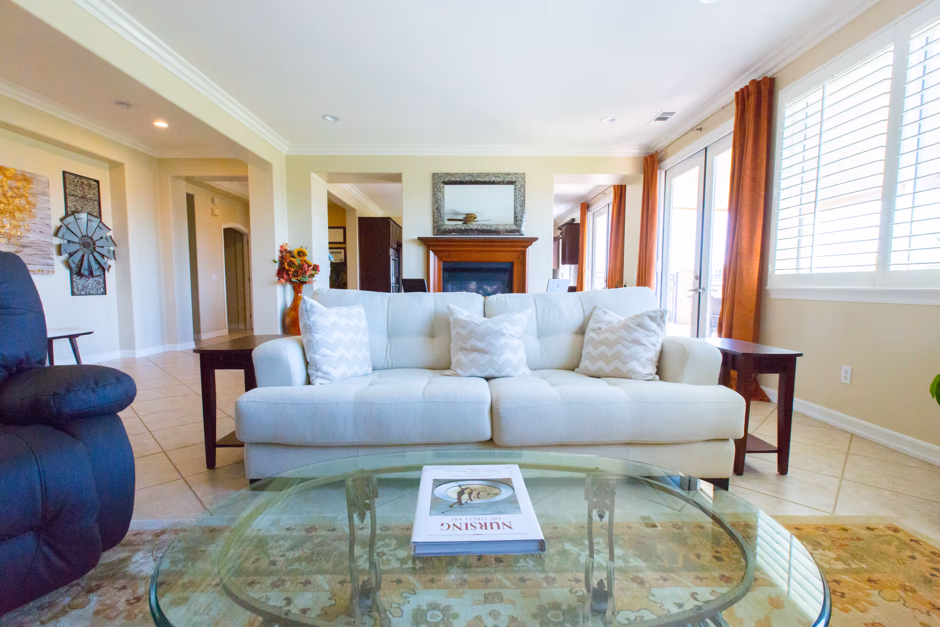 Bright living room with a white couch adorned with three chevron-patterned pillows, a glass coffee table with a book titled 'Nursing The Finest Art', a dark recliner chair on the left, a fireplace with a mirror above it in the background, and large windows with white blinds and orange curtains letting in natural light.