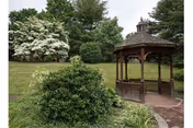 A wooden gazebo beside a paved walkway surrounded by bushes, a grassy lawn, and trees.
