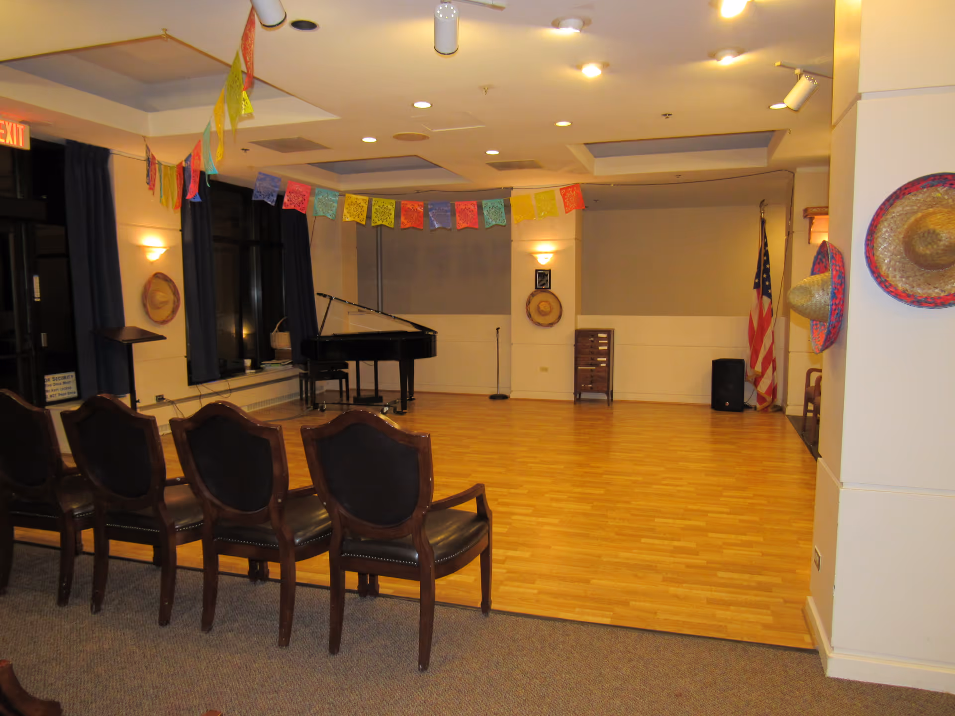 Empty community activity room with rows of chairs facing a grand piano on a wood floor, decorated with colorful banners and sombreros.