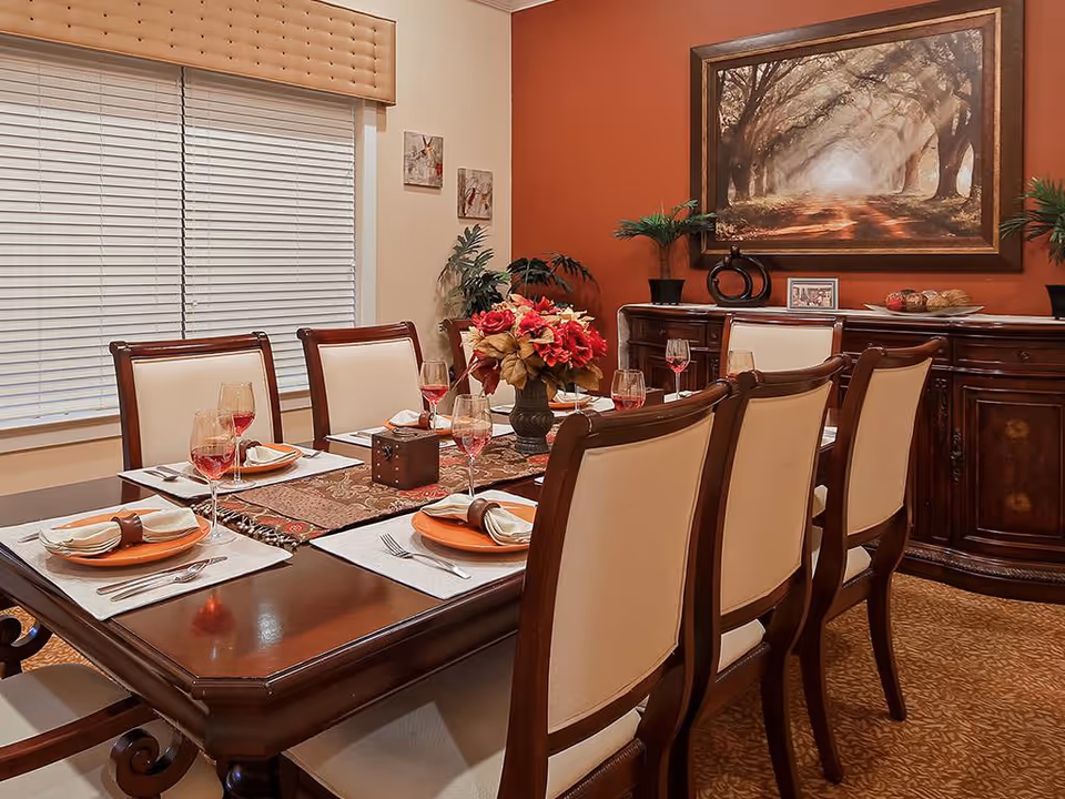Elegant dining room with a set wooden table, upholstered chairs, floral centerpiece and sideboard beneath a framed painting.