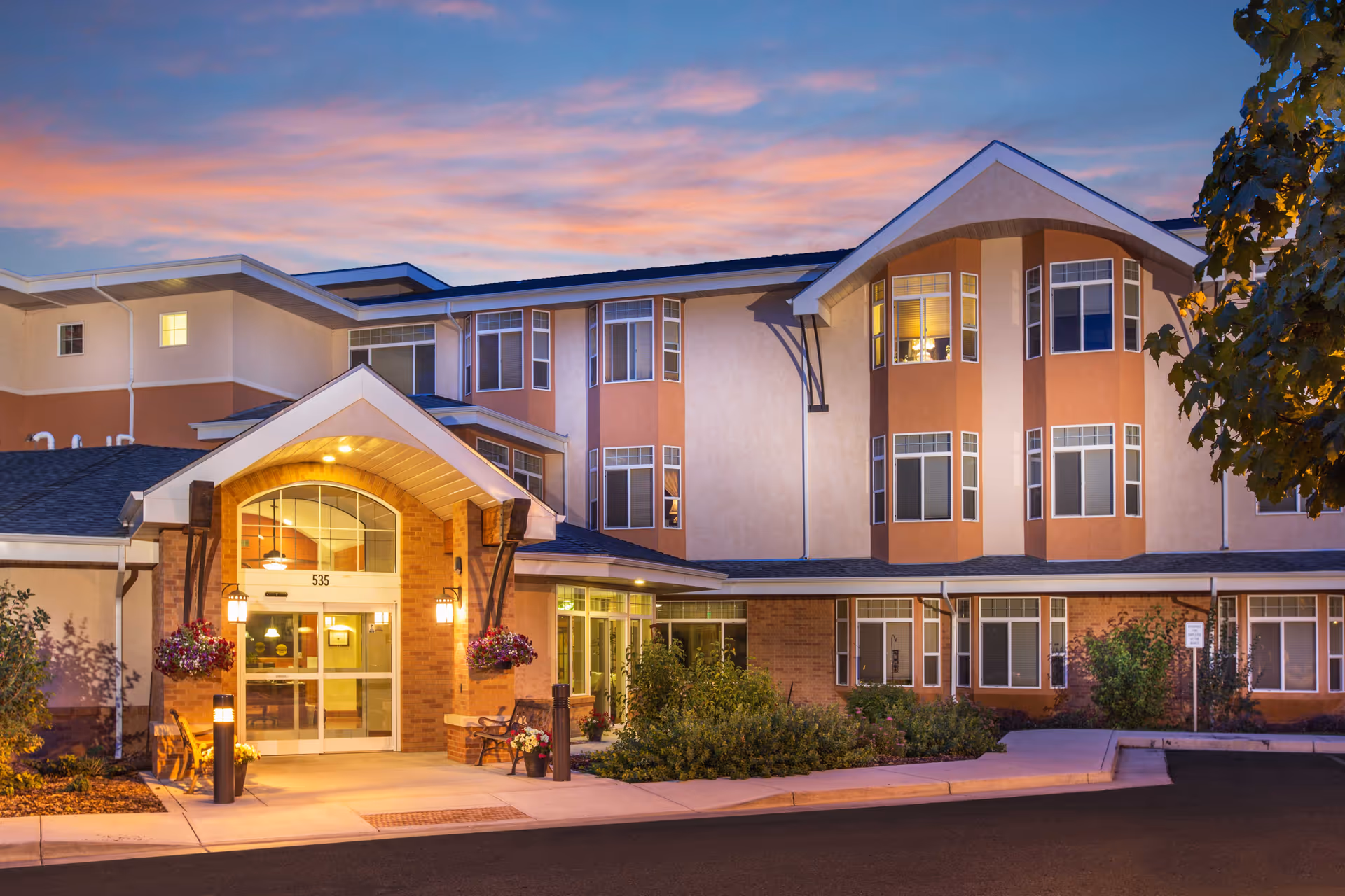 Exterior view of Hillcrest Of Loveland senior living facility at dusk, showing the entrance with warm lighting, hanging flower baskets, and a well-maintained garden area under a colorful sky.
