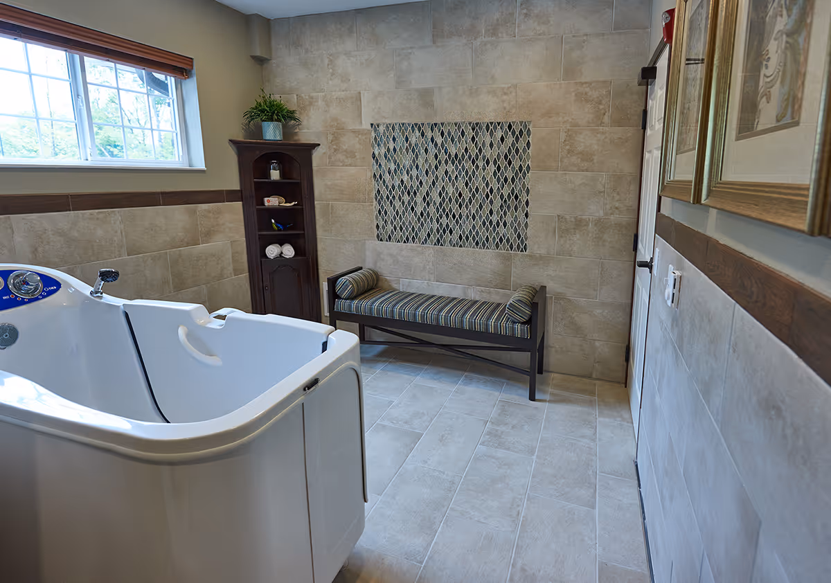 A tiled bathroom with a walk-in bathtub on the left, a wooden shelving unit with towels and toiletries, a cushioned bench with striped upholstery against the wall, and framed artwork on the right wall. There is a window with wooden blinds letting in natural light.