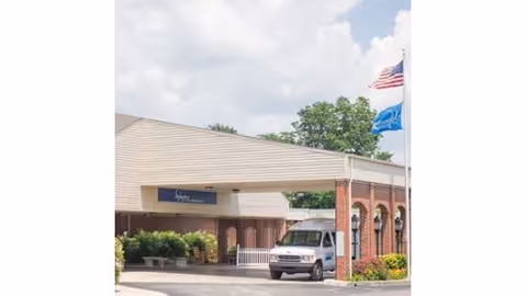 Covered entrance/porte-cochere of a senior care facility with a white van parked underneath and flags flying nearby.