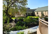 Outdoor garden area with a curved walkway and railing, surrounded by green plants and trees, adjacent to a brick building under a clear blue sky.