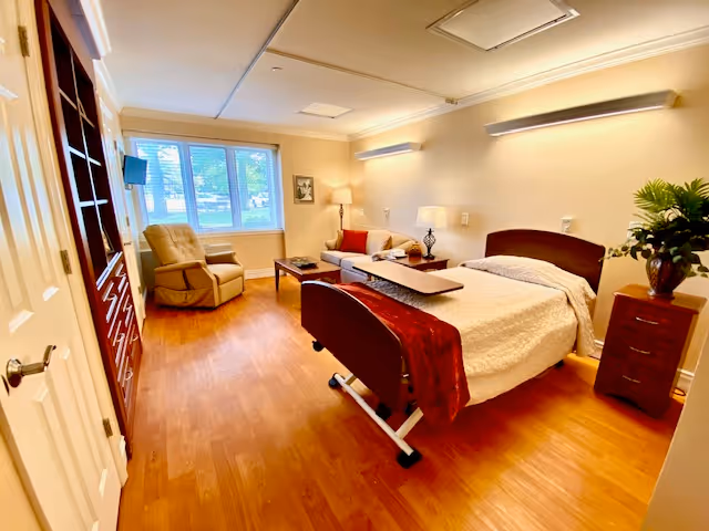 A well-lit senior living bedroom featuring a hospital-style adjustable bed with white bedding and a red throw blanket. The room includes a wooden nightstand with a plant, a beige sofa with red cushions, a coffee table, a beige recliner chair near a window with blinds, and a wall-mounted TV. The floor is wooden, and the walls are painted a light color with modern lighting fixtures.
