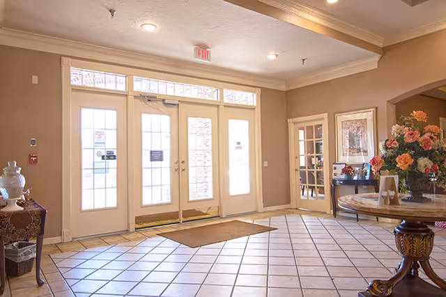Bright and spacious entrance lobby of a senior living facility with large glass double doors, tiled floor, a round wooden table with a floral arrangement, and framed artwork on the walls.