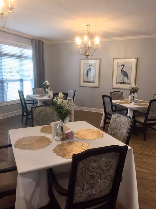 A dining room with several tables covered in white tablecloths and gold placemats. Each table has a small floral centerpiece with white flowers. The room has wooden flooring, light gray walls, and two framed bird illustrations on the wall. A chandelier with lit bulbs hangs from the ceiling, and a large window with blinds and gray curtains lets in natural light.