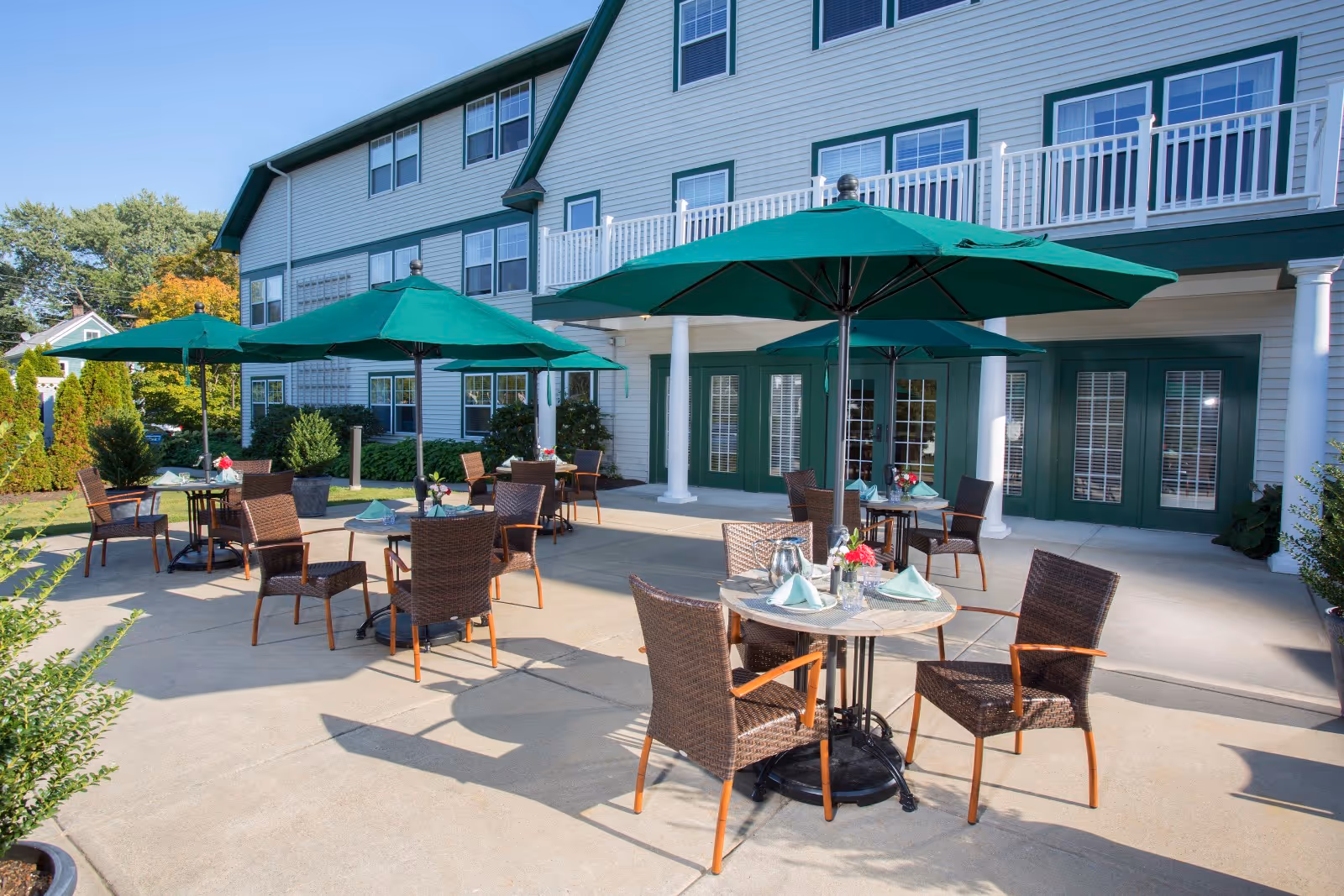 Outdoor patio area at Crescent Point at Niantic with several round tables, each set with napkins, glasses, and small flower arrangements. The tables are shaded by large green umbrellas, and wicker chairs surround each table. The patio is adjacent to a multi-story building with white siding and green trim, featuring multiple windows and doors.