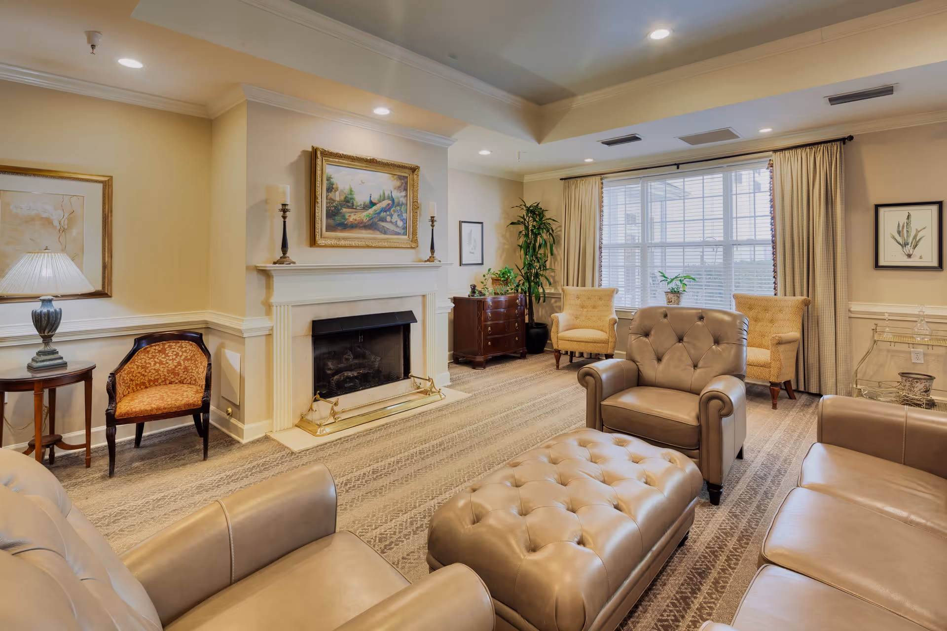 A cozy living room with beige walls and carpeted floor featuring a fireplace with a painting above it. The room is furnished with leather sofas, an armchair, a tufted ottoman, two upholstered chairs near a large window with curtains, a side table with a lamp, a wooden chest, and some potted plants.