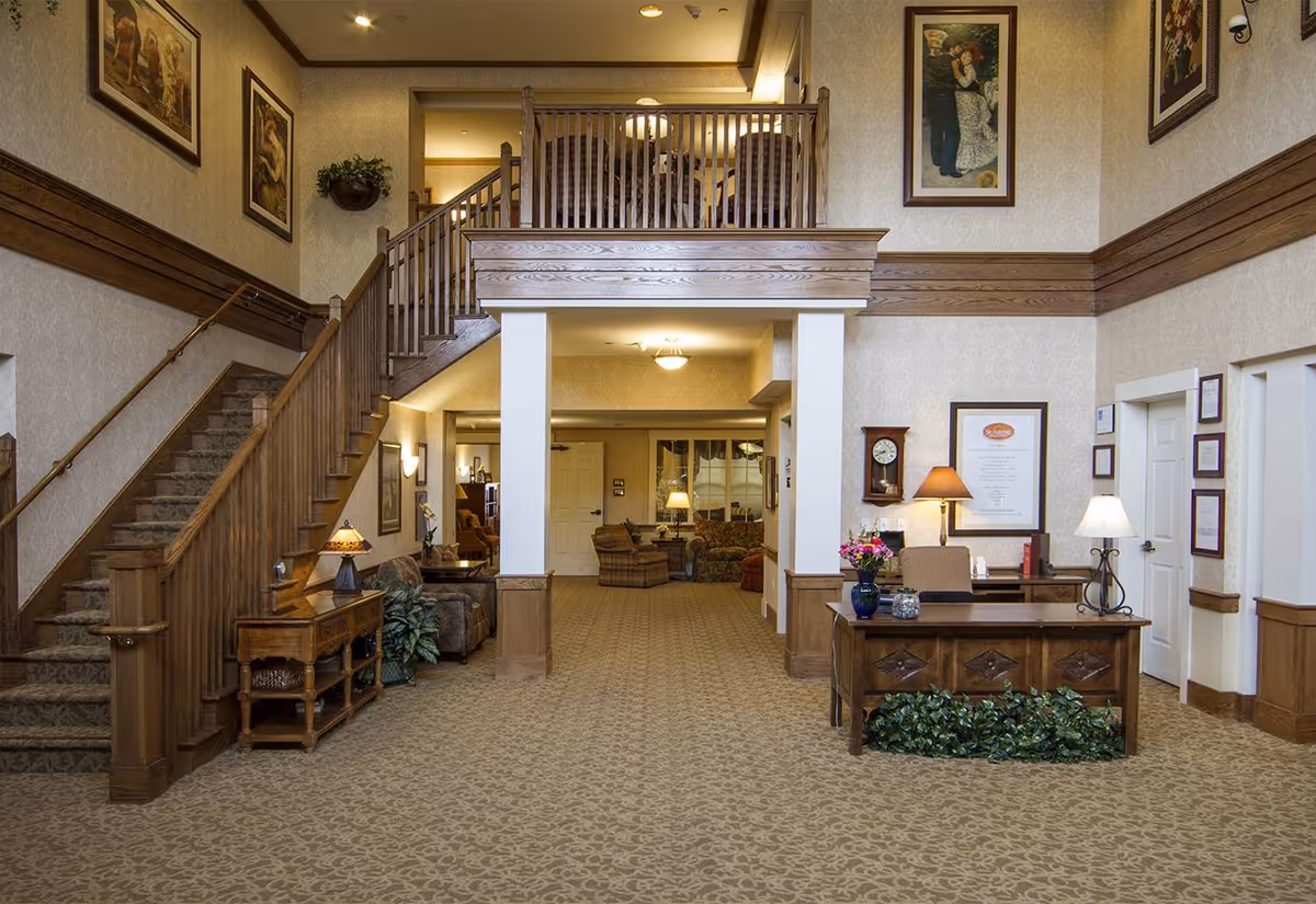 A spacious, warmly decorated senior living facility lobby with a wooden reception desk, staircase, and seating area.