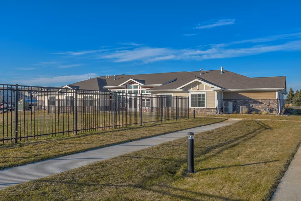 Exterior view of a single-story senior living facility with a black metal fence, walkway, and manicured lawn under a blue sky.