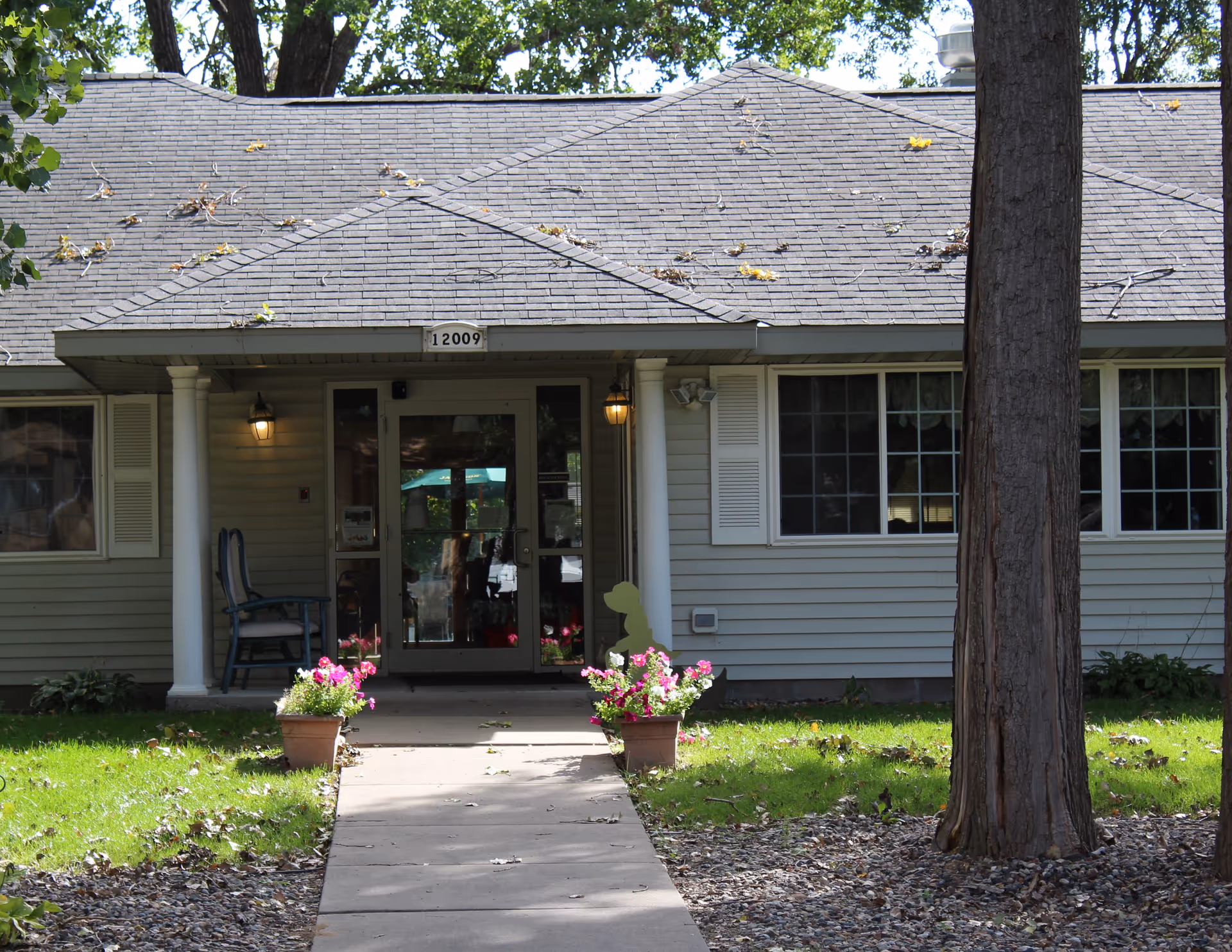Front entrance of a single-story building with a gray shingled roof and light gray siding. The entrance has double glass doors with two white columns on either side and two wall-mounted lantern lights. There are two flower pots with pink and white flowers on either side of the concrete walkway leading to the door. A blue chair is placed on the left side porch area. Trees and green grass surround the building.