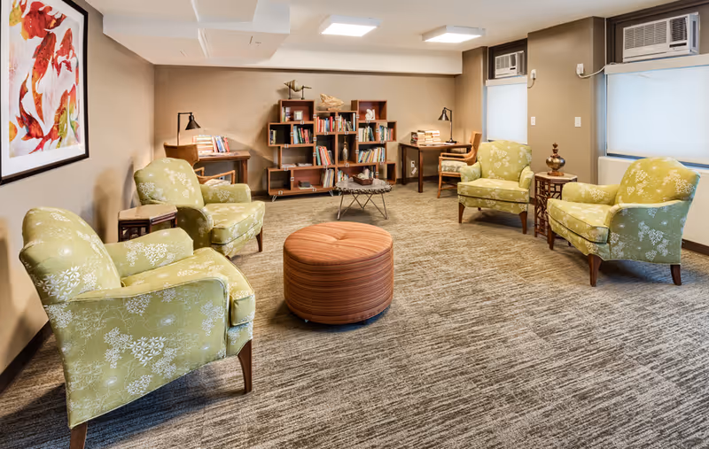 A cozy living room area with four green patterned armchairs arranged around a round orange ottoman. The room features a beige carpet, two desks with chairs and lamps against the back wall, a bookshelf filled with books and decorative items, two windows with air conditioning units, and a large colorful abstract painting on the left wall.